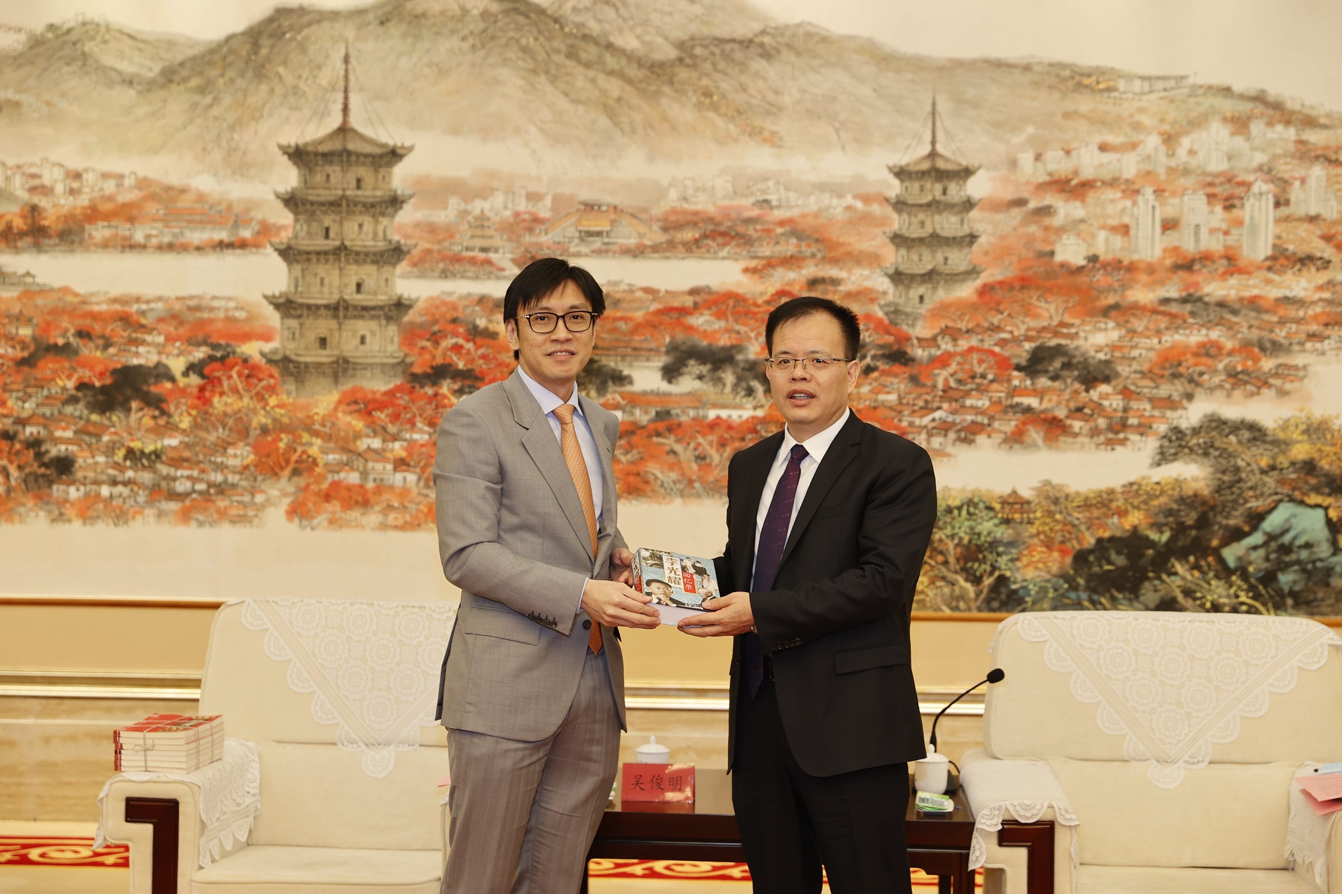 Two men in suits exchanging a book in front of a landscape painting.